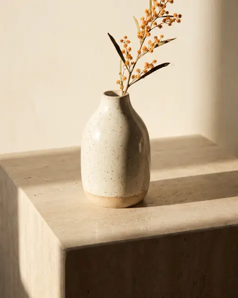 Speckled stoneware bud vase with a dried wattle stem on a travertine plinth